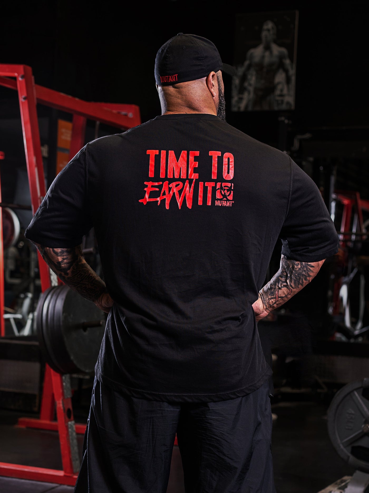  Back view of Dusty Hanshaw, Mutant athlete, posing in the gym wearing the Black 'Dusty Says' Mutant Oversized Gym T-shirt with  the phrase 'Time to earn it' and the Mutant logo in red.