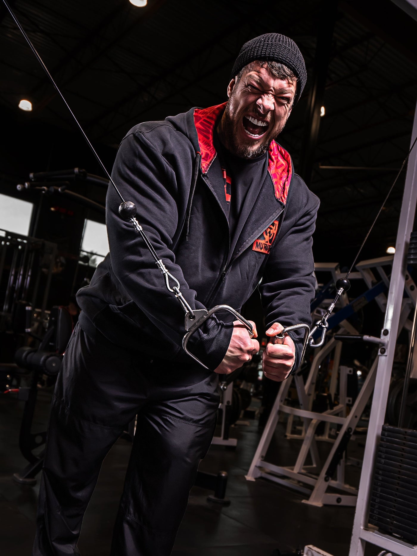Mutant athlete Jamie Christian is training in the gym wearing Black Mutant Patched Zip-Up Gym Hoodie with a red and black Mutant logo patch on left chest. Two pockets and red hood lining with red watermarked Mutant logo