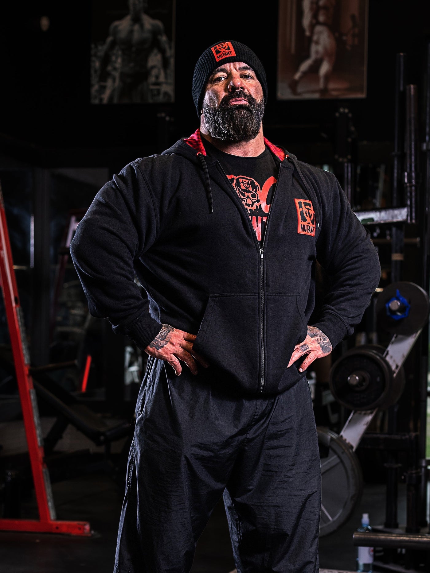 Mutant athlete Dusty Hanshaw posing in the gym wearing Black Mutant Patched Zip-Up Gym Hoodie with a red and black Mutant logo patch on left chest. Two pockets and red hood lining with red watermarked Mutant logo.