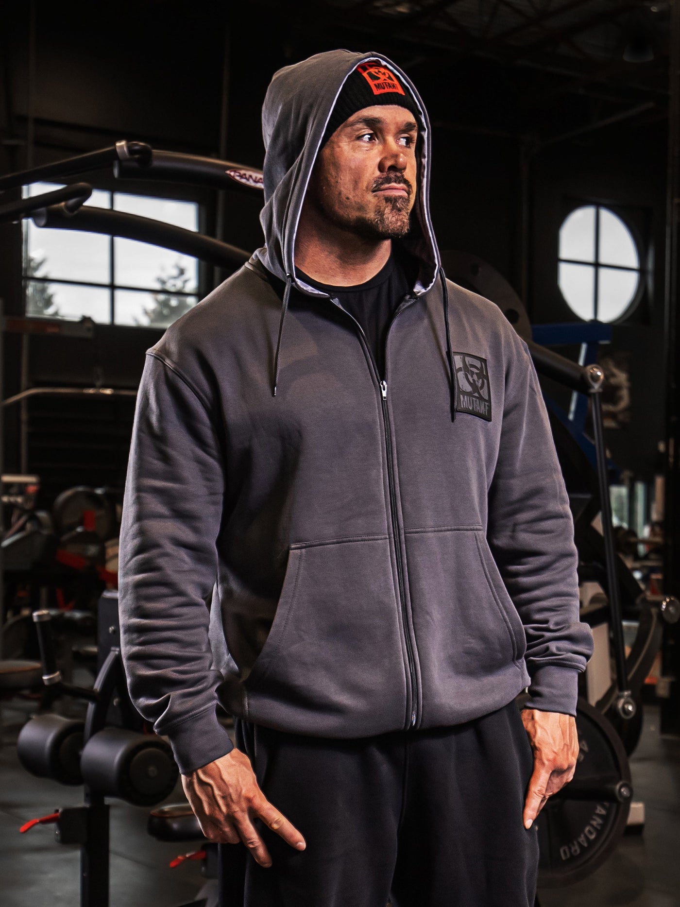 Mutant athlete Ron Partlow posing in the gym, wearing the Grey Mutant Patched Zip-Up Gym Hoodie. The hoodie features a black Mutant logo patch on the left chest, two pockets, and a light grey hood lining with a watermarked Mutant logo.