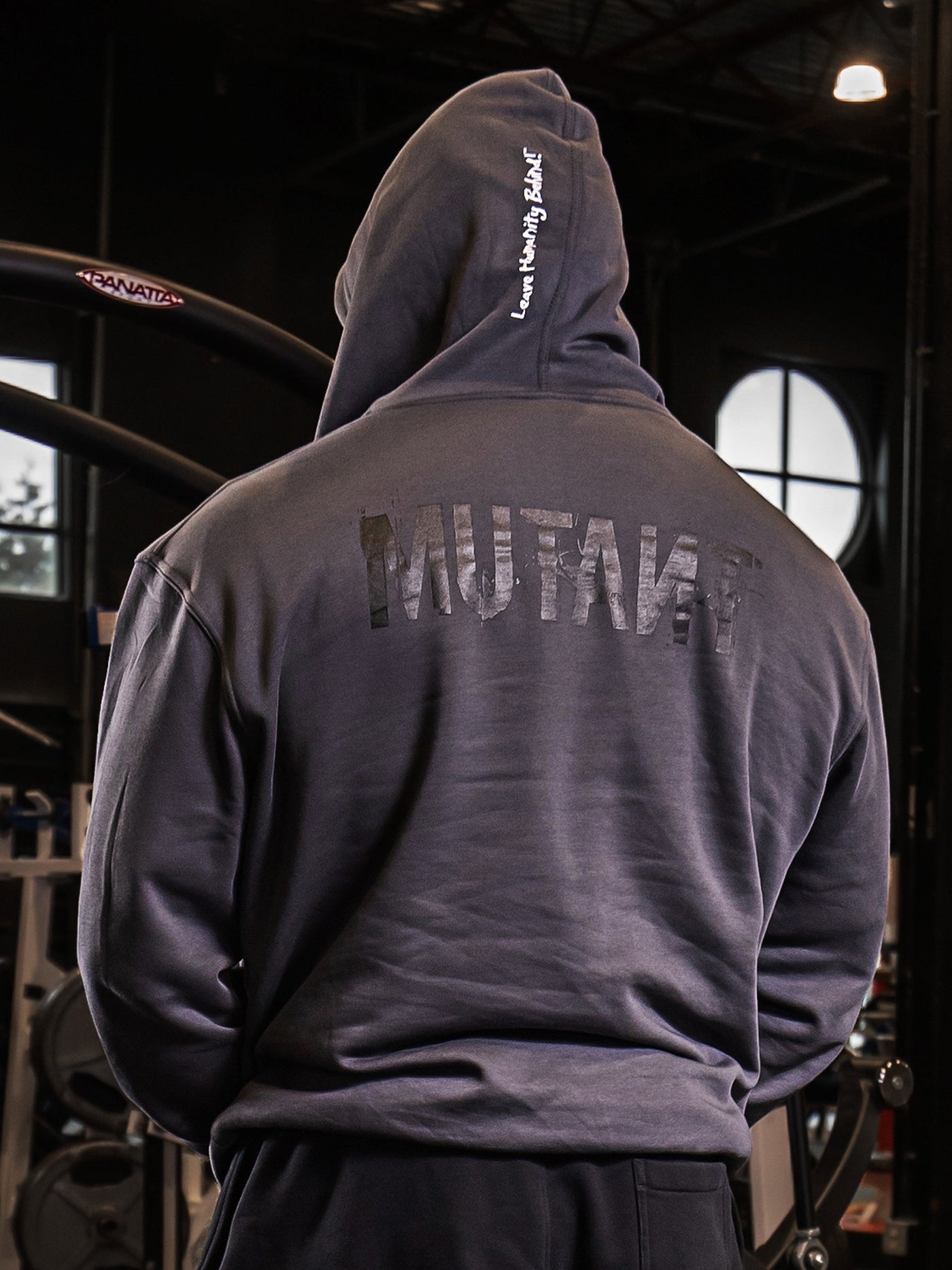 Back view of an athlete posing in the gym, wearing the Grey Mutant Patched Zip-Up Gym Hoodie. The hoodie features a black Mutant logo on the back and the white Mutant motto 'Leave Humanity Behind' on the hood.
