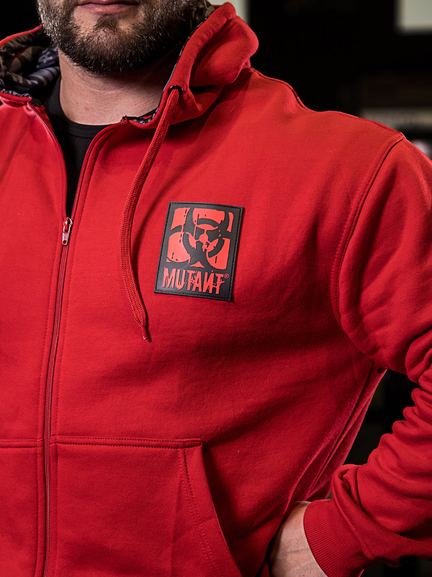 Close-up photo of a Mutant athlete wearing the red Mutant Patched Zip-Up Gym Hoodie. The image focuses on the black and red Mutant logo patch on the left chest, showcasing the intricate details.