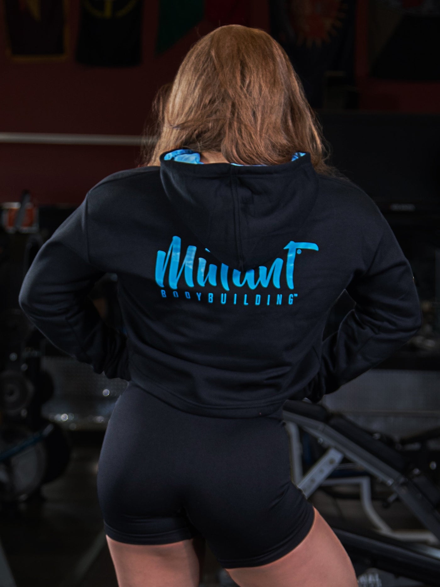 Shelby Guillaume, Mutant athlete, posing in the gym to showcase the back of the black Mutant Thick Script Women's Gym Crop Hoodie.